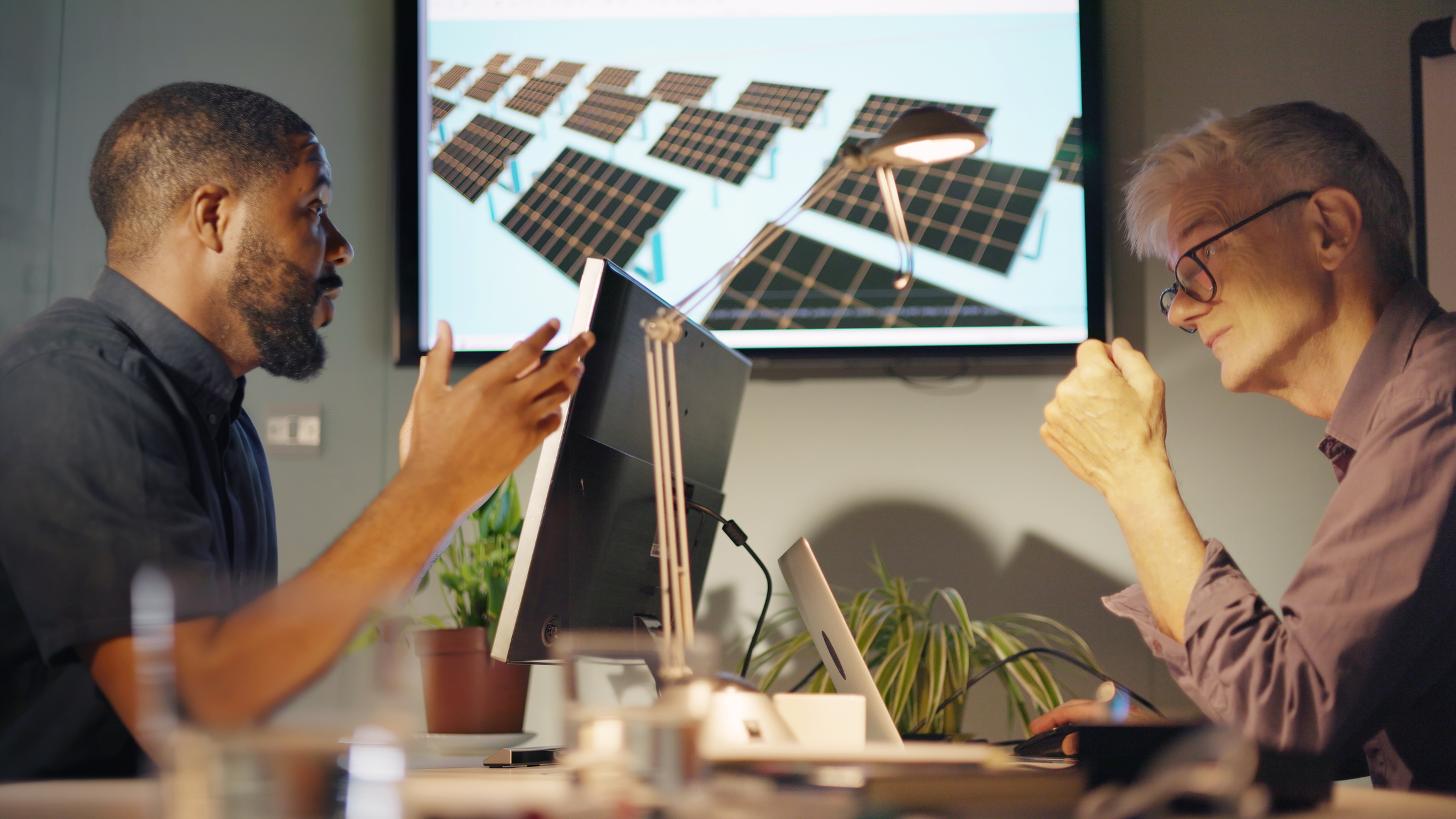 Stock image of two men, one black and the other white &amp; mature meeting to discuss solar pale installation design… this is shown on a screen in the office.
