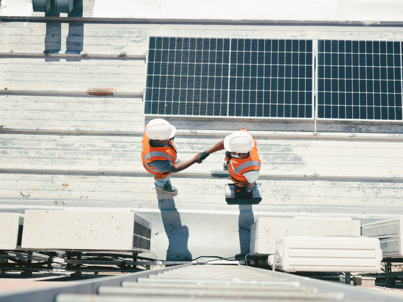 Two construction workers in orange vests and white helmets, standing on a rooftop next to solar panels, shake hands. They are beside HVAC units and appear to be collaborating on the renewable energy rooftop project. The view is from above.